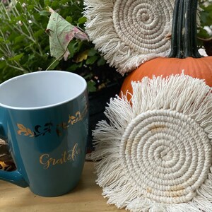 K&ouml;nnte beinhalten: Zwei wei&szlig;e geh&auml;kelte Untersetzer mit Fransenrand. Die Untersetzer sind rund und haben ein Spiraldesign. Die Untersetzer liegen auf einer Holzoberfl&auml;che neben einer t&uuml;rkisfarbenen Tasse mit dem Wort "Grateful" in Gold.