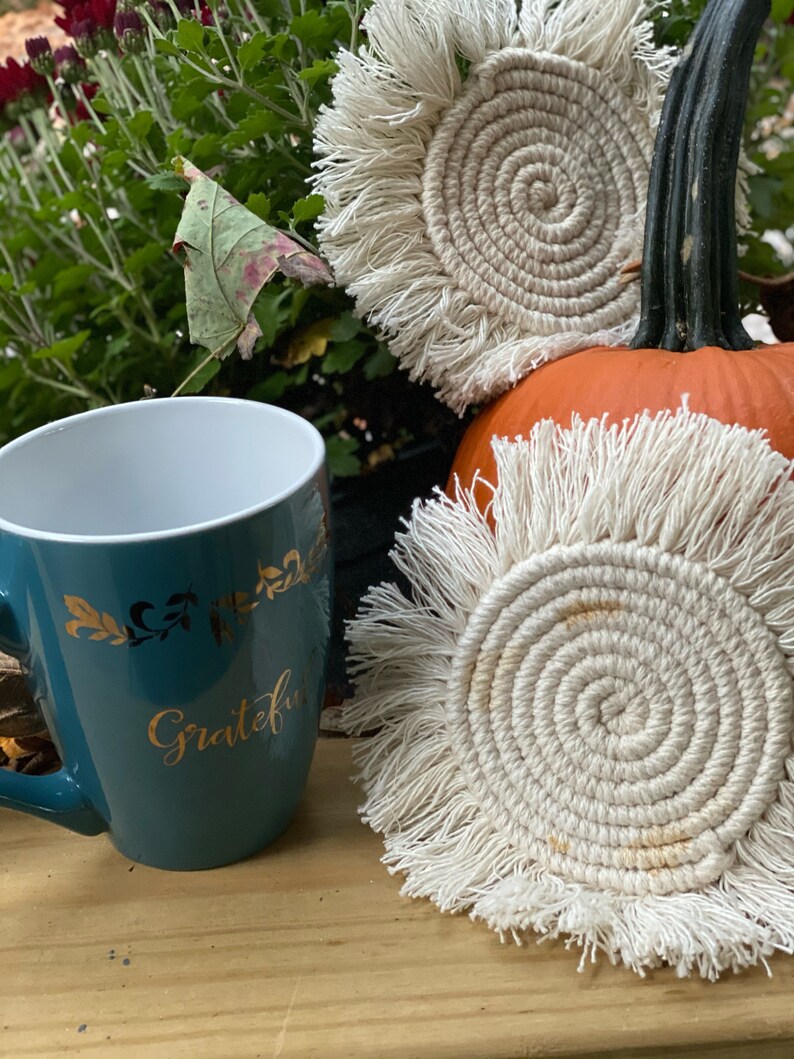 K&ouml;nnte beinhalten: Zwei wei&szlig;e geh&auml;kelte Untersetzer mit Fransenrand. Die Untersetzer sind rund und haben ein Spiraldesign. Die Untersetzer liegen auf einer Holzoberfl&auml;che neben einer t&uuml;rkisfarbenen Tasse mit dem Wort "Grateful" in Gold.