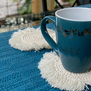 K&ouml;nnte beinhalten: Eine t&uuml;rkisfarbene Tasse mit goldenen Blattdetails und dem Wort "grateful" auf der Seite, die auf einem wei&szlig;en geh&auml;kelten Untersetzer mit Fransen steht. Der Untersetzer befindet sich auf einer blauen gewebten Tischdecke.