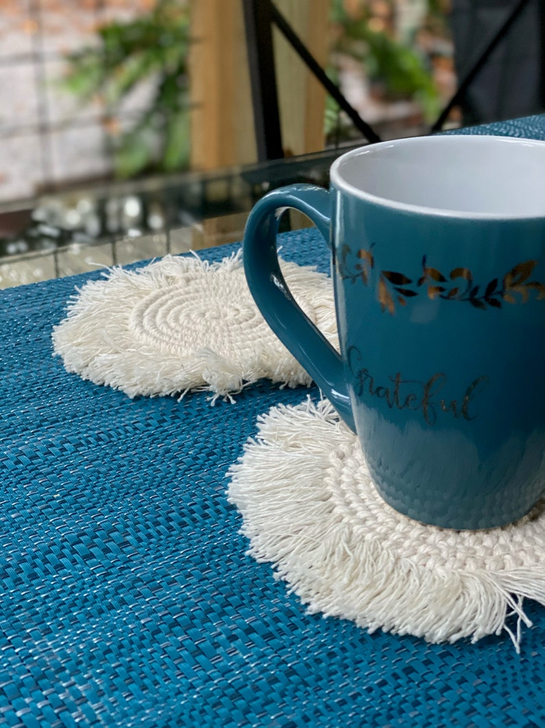 K&ouml;nnte beinhalten: Eine t&uuml;rkisfarbene Tasse mit goldenen Blattdetails und dem Wort "grateful" auf der Seite, die auf einem wei&szlig;en geh&auml;kelten Untersetzer mit Fransen steht. Der Untersetzer befindet sich auf einer blauen gewebten Tischdecke.