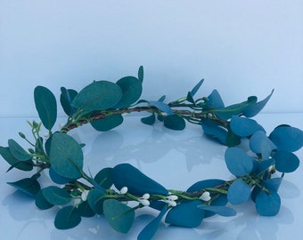 May include: A circular floral crown featuring blue-green eucalyptus leaves and small white berries. The leaves are arranged around a brown, twig-like base, creating a natural and delicate aesthetic. This headpiece is suitable for weddings, festivals, or everyday wear.
