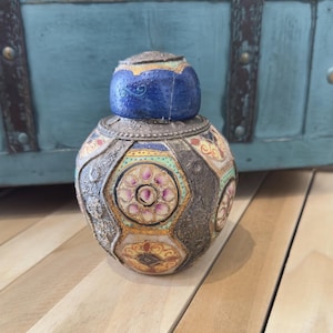 Op de afbeelding: Een decoratieve fles in antieke stijl met een blauwe dop. De fles heeft een zilverkleurige behuizing met kleurrijke, geometrische patronen in geel, roze en turquoise. Hij staat op een houten oppervlak, met een blauwe kist op de achtergrond.