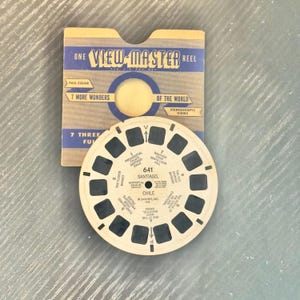 May include: A vintage View-Master reel and its original cardboard sleeve. The beige circular reel features seven pairs of image frames labeled with Santiago, Chile landmarks. The blue and tan sleeve displays the text One View-Master Reel.