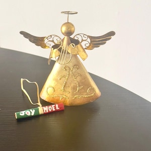 Può includere: Ciondolo a forma di angelo in metallo dorato con aureola, ali e arpa. L'angelo tiene un piccolo ciondolo rosso e verde che dice "Joy Noel".