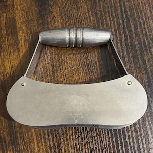 May include: A silver metal garlic press with a curved handle. The handle has a textured grip. The press has a small logo on the front that says "Garlic Press" and "Made in USA".