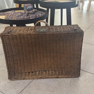 Puede incluir: Una maleta rectangular de mimbre tejido con asa y cierre metálico. La maleta es de color marrón cálido y parece ser vintage. El mimbre está tejido apretado y la maleta tiene forma rectangular.