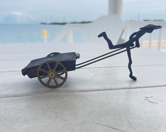 Peut inclure: Une petite sculpture en métal d'une personne tirant un chariot à une seule roue. La sculpture est en métal noir et a une roue dorée. La sculpture est sur une surface en bois blanc.