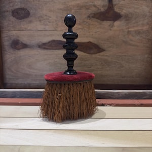 Puede incluir: Un cepillo pequeño con mango negro y base de terciopelo rojo. El cepillo tiene cerdas naturales y es ideal para limpiar superficies delicadas.