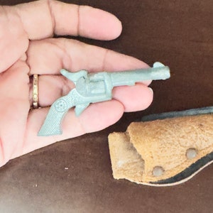 Puede incluir: Un revólver de juguete plateado con una funda de cuero marrón. La pistola es pequeña y tiene un diseño sencillo.