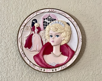 May include: A pink and gold ceramic plate with a raised image of a blonde Barbie doll wearing a red dress and a pearl necklace. The plate has a gold trim and the word "Barbie" written in gold letters.