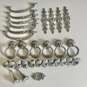May include: A collection of silver-toned drawer pulls and cabinet knobs. The set includes a variety of styles, including ring pulls, ornate pulls, and decorative knobs.