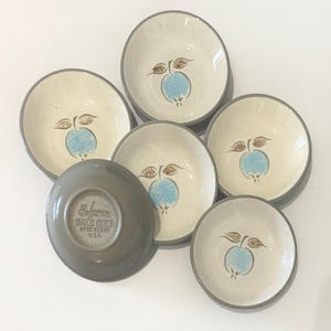 Puede incluir: Un conjunto de seis cuencos de gres vintage. Los cuencos tienen un interior color crema con un diseño de manzana azul y hojas marrones. Los bordes son grises. Un cuenco está boca abajo, revelando una base gris con texto.