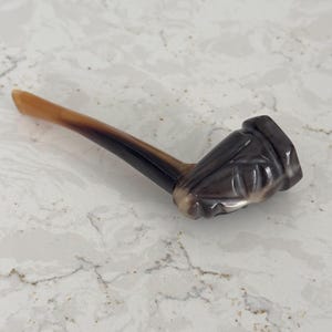 May include: A smoking pipe crafted from polished horn, featuring a smooth, curved stem that transitions from a light tan to a deep, dark brown. The bowl is intricately carved with a stylized face, showcasing natural grey and white marbling.