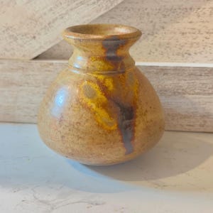 May include: A small, round ceramic vase with a wide, bulbous body and a narrow neck. The surface features a mottled, earthy tan glaze with prominent streaks of deep brown and vibrant yellow running vertically down the side of the vessel.