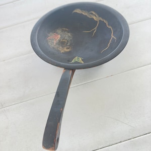 May include: A black wooden bowl with a long handle. The bowl has a painted design of a branch with leaves and a circle with a swirl pattern.
