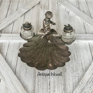 May include: Antique silver inkwell with a cherub figurine and two glass inkwells. The inkwell is shaped like a seashell and has a decorative design.
