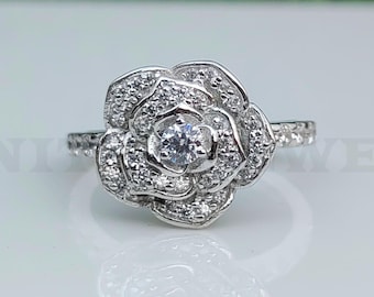 May include: A silver ring featuring a rose-shaped design. The ring's center showcases a round, clear gemstone, surrounded by smaller stones that form the petals. The band is also adorned with small stones, adding to the ring's sparkle.