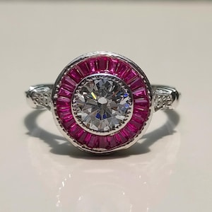 May include: A silver ring with a central, clear gemstone surrounded by a ring of smaller, rectangular, pink gemstones. The ring's band has decorative detailing on the sides. The ring is set against a white surface with a blurred green background.