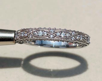 May include: A silver ring featuring an ornate filigree pattern and small, clear gemstones. The ring is held by a metal tool, highlighting its detailed design and sparkling stones. The ring's shadow is cast on a light surface.