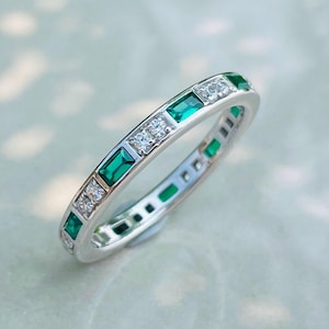 May include: A silver ring with alternating emerald-cut green stones and small, round, clear stones. The ring has a continuous pattern of gemstones around the band. The ring is set on a light-colored surface.