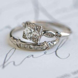 May include: A silver-toned engagement ring set featuring a central round-cut stone in a square setting, flanked by small accent stones on the band, paired with a matching wedding band adorned with a row of five small round-cut stones.