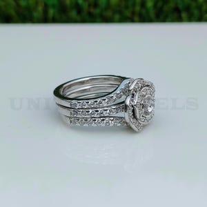 May include: A three-piece silver ring set. The engagement ring features a large, round center stone surrounded by smaller stones. The bands are also adorned with small, sparkling stones. The rings are displayed on a white surface.