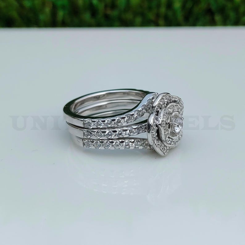 May include: A three-piece silver ring set. The engagement ring features a large, round center stone surrounded by smaller stones. The bands are also adorned with small, sparkling stones. The rings are displayed on a white surface.