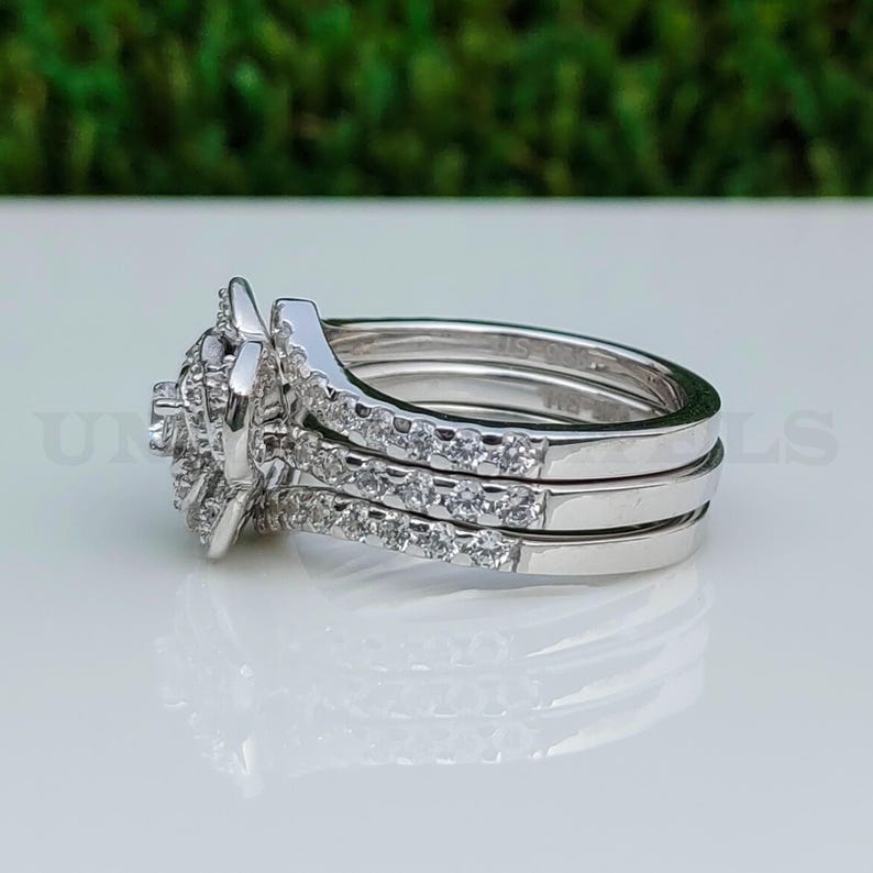 May include: A three-piece silver ring set. The engagement ring features a central cluster of small, round stones. Two additional bands are encrusted with smaller stones. The rings are displayed on a white surface with a blurred green background.