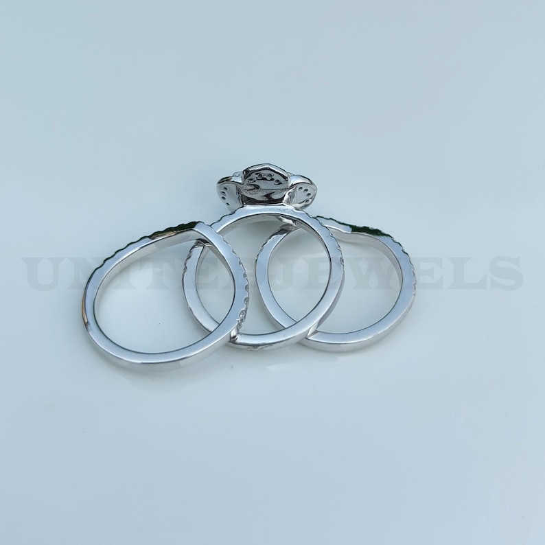 May include: A set of three silver rings. The engagement ring features a central stone surrounded by smaller stones. The other two rings have a band of small stones. The rings are displayed on a light background.