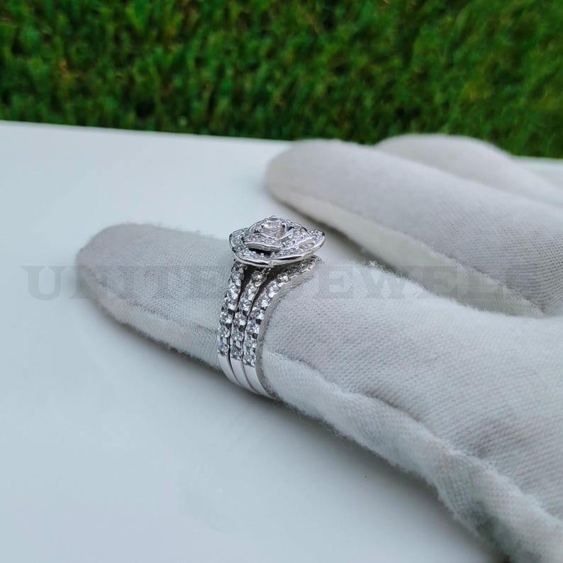 May include: A three-piece silver ring set. The engagement ring features a central, round-cut diamond surrounded by a halo of smaller stones. Two additional bands, also adorned with small diamonds, complete the set. The rings are displayed on a white surface.