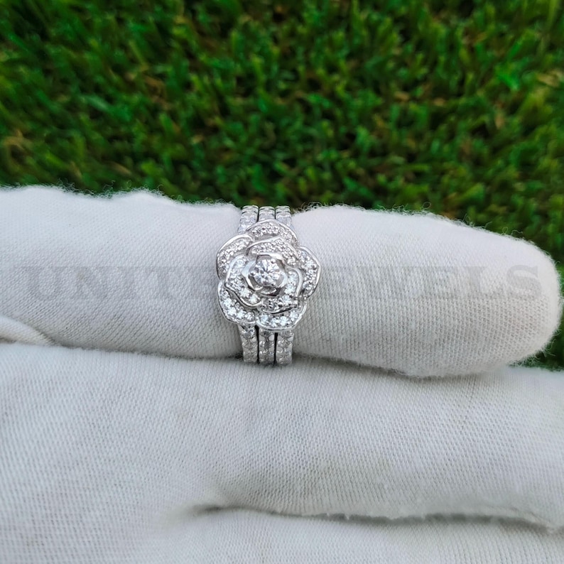 May include: A silver ring set featuring a floral design. The ring has a central flower shape with multiple small stones and a triple-band design. The ring is displayed on a white surface against a green background.