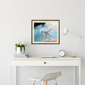 May include: A framed painting of a white dog running through a blue and white abstract landscape. The painting is hanging on a white wall above a white desk with a white chair.