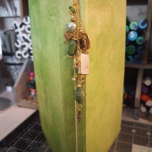 May include: A green book with a gold chain bookmark. The bookmark has green and gold beads, a small gold charm, and a white paper bead.
