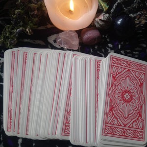 May include: A deck of tarot cards with red patterned backs is fanned out in the foreground. A lit white candle, crystals, and other spiritual items are arranged in the background. The cards have white faces and red borders.