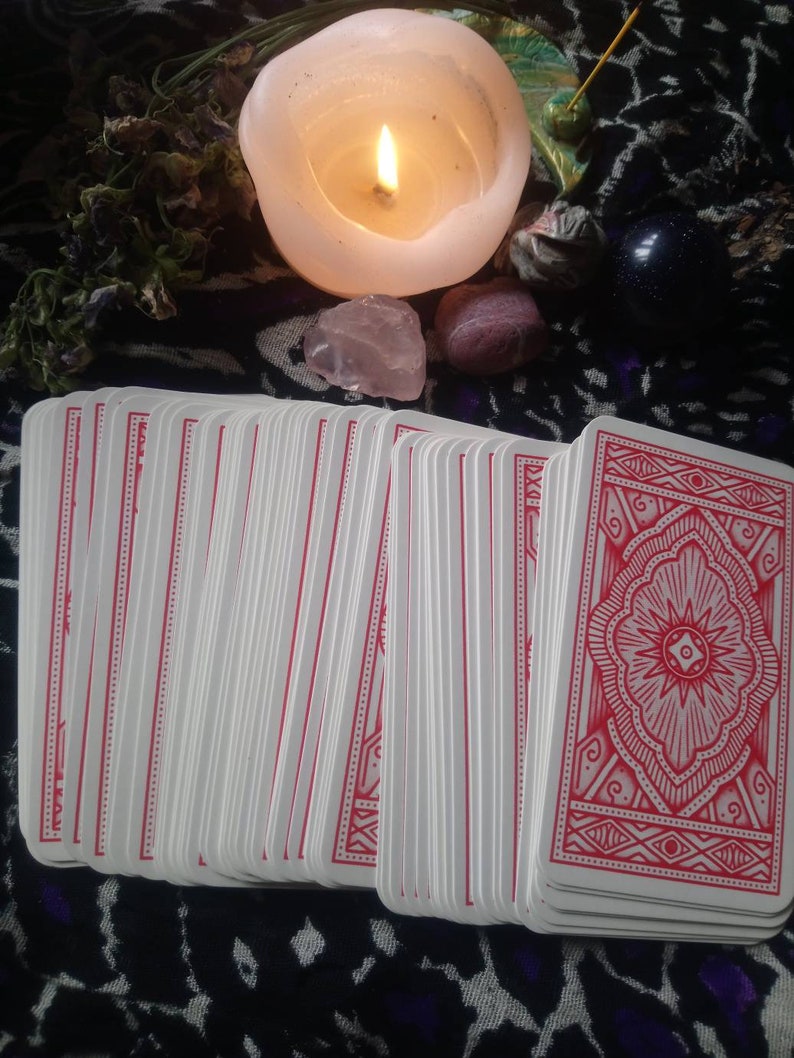 May include: A deck of tarot cards with red patterned backs is fanned out in the foreground. A lit white candle, crystals, and other spiritual items are arranged in the background. The cards have white faces and red borders.