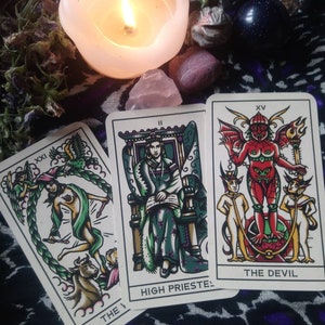 May include: Three tarot cards are displayed, featuring illustrations and text. The cards are placed on a patterned surface, with a lit candle, crystals, and dried flowers in the background. The cards are labeled "The World", "High Priestess", and "The Devil".