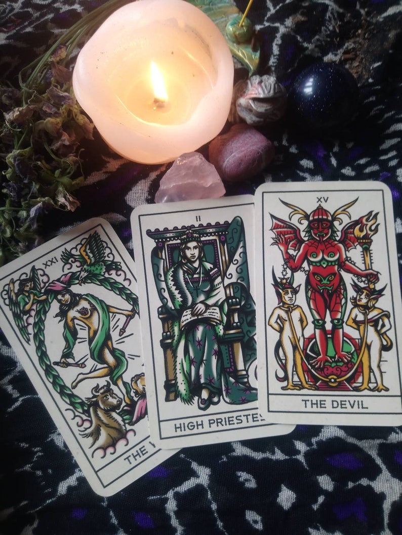 May include: Three tarot cards are displayed, featuring illustrations and text. The cards are placed on a patterned surface, with a lit candle, crystals, and dried flowers in the background. The cards are labeled "The World", "High Priestess", and "The Devil".