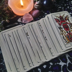 May include: A lit candle, tarot cards, and crystals are arranged on a patterned cloth. The tarot cards are fanned out, with the top card showing a red devil figure. A pink candle and crystals are also visible.