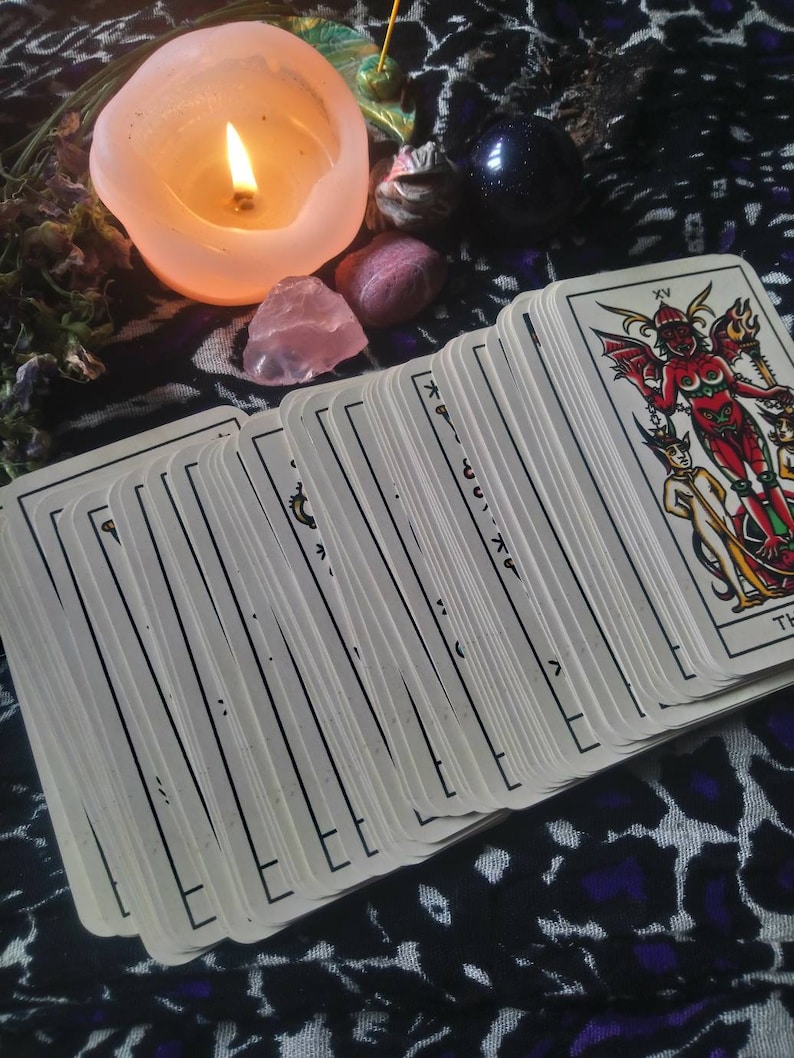 May include: A lit candle, tarot cards, and crystals are arranged on a patterned cloth. The tarot cards are fanned out, with the top card showing a red devil figure. A pink candle and crystals are also visible.