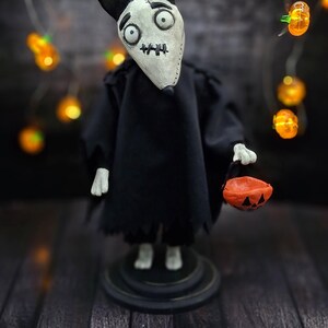 Handmade Sparky Art Doll wearing a black hoodie Halloween-style outfit, holding a pumpkin-shaped trick-or-treat box. Standing straight, looking surprised directly at the viewer. Whimsical and spooky Halloween home decor, unique collectible dog doll