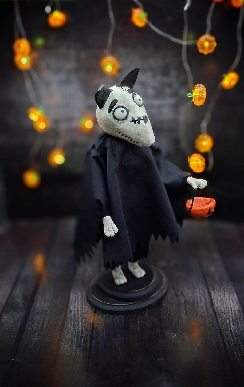 Handmade Sparky Art Doll wearing a black hoodie Halloween-style outfit, holding a pumpkin-shaped trick-or-treat box. Shown from a right diagonal angle. Whimsical and spooky Halloween home decor, unique collectible dog doll