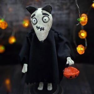 Handmade Sparky Art Doll wearing a black hoodie Halloween-style outfit, holding a pumpkin-shaped trick-or-treat box. Standing straight, looking down and to the left. Whimsical and spooky Halloween home decor, unique collectible dog doll