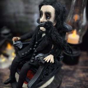 Profile view from the left of a handmade Edgar Allan Poe art doll sitting on a rocking chair on a small carpeted stand, with a raven in the foreground, a poseable art doll and unique Poe collectible perfect for literary decor, gothic home accent, and dark fantasy displays, highlighting detailed sculpting and painted features.