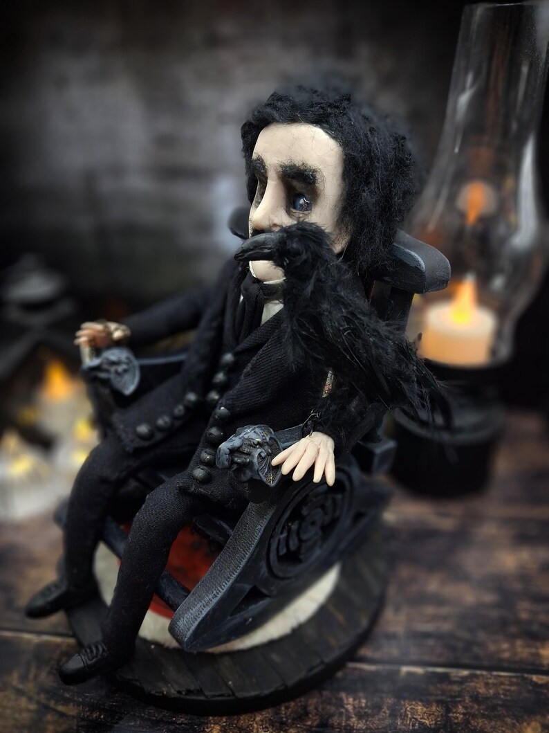 Profile view from the left of a handmade Edgar Allan Poe art doll sitting on a rocking chair on a small carpeted stand, with a raven in the foreground, a poseable art doll and unique Poe collectible perfect for literary decor, gothic home accent, and dark fantasy displays, highlighting detailed sculpting and painted features.