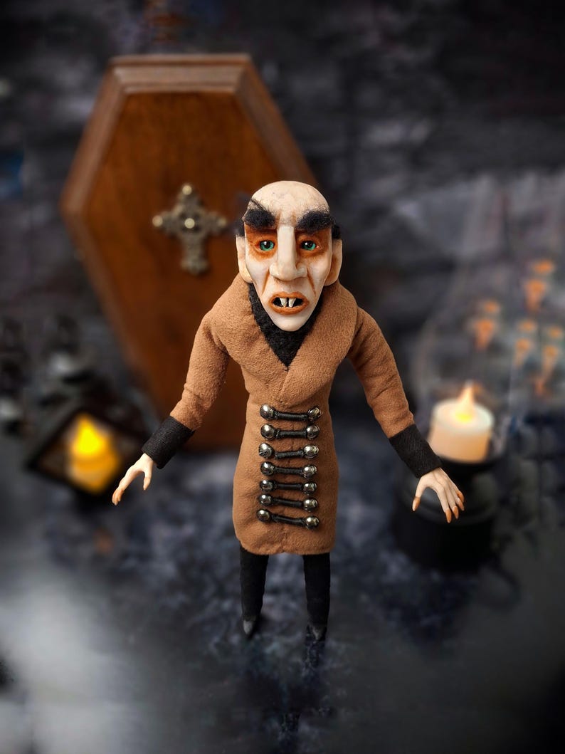Wide front view of a handmade Nosferatu art doll standing in front of a closed wooden coffin, designed with special fabrics and accessories, featuring a cross on top, a unique gothic vampire collectible perfect for spooky art displays, gothic decor, horror themed collections, and dark fantasy setups, highlighting detailed sculpting and costume features.