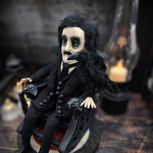 Front-left diagonal view of a handmade Edgar Allan Poe art doll sitting on a rocking chair on a small carpeted stand, with a raven perched nearby, a poseable art doll and unique Poe collectible perfect for literary decor, gothic home accent, and dark fantasy displays, highlighting detailed sculpting and painted features.