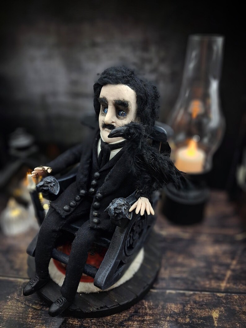 Front-left diagonal view of a handmade Edgar Allan Poe art doll sitting on a rocking chair on a small carpeted stand, with a raven perched nearby, a poseable art doll and unique Poe collectible perfect for literary decor, gothic home accent, and dark fantasy displays, highlighting detailed sculpting and painted features.
