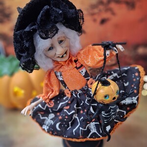 Top right angled view of the handmade witch art doll holding a tiny pumpkin skeleton puppet, showing her detailed Halloween outfit and whimsical spooky design.
