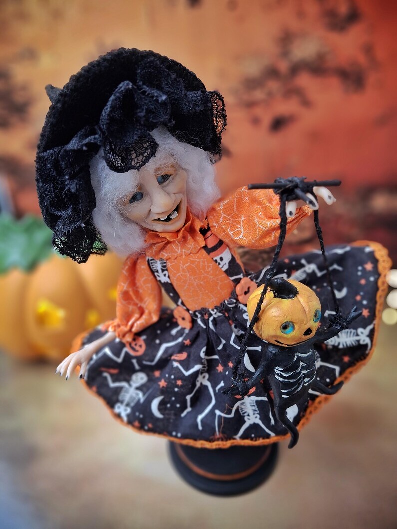 Top right angled view of the handmade witch art doll holding a tiny pumpkin skeleton puppet, showing her detailed Halloween outfit and whimsical spooky design.