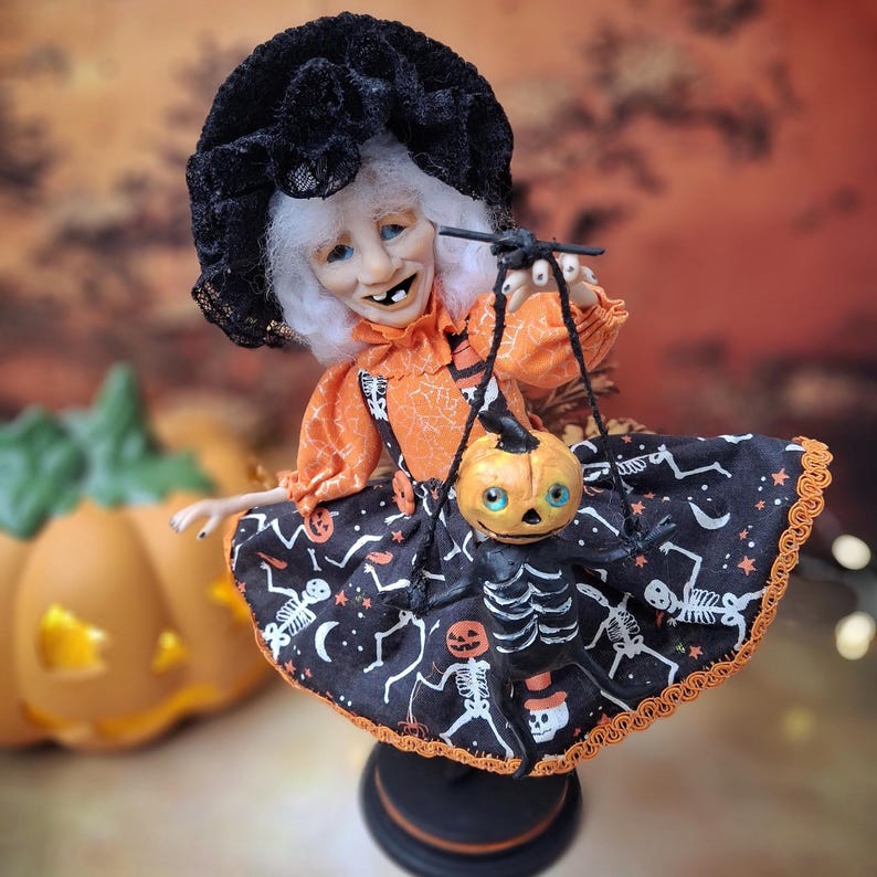 A handmade witch art doll standing upright, holding a tiny handmade pumpkin skeleton puppet in one hand. She wears an adorable Halloween-themed outfit with spooky cute details, perfect for autumn and Halloween decor.